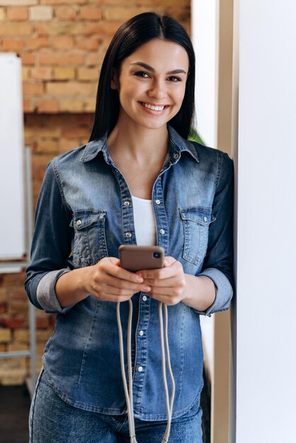 Молодая брюнетка дама в джинсовой одежде держит смартфон и счастливо улыбается в камеру, стоя в интерьере офиса