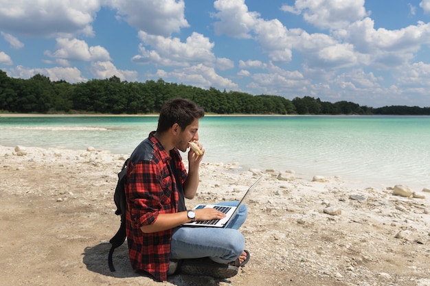Young brunette male in jeans and shirt is working at a computer while being on the nature. 