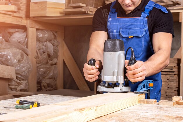 A young brunette man carpenter equals a wooden plank with a milling machine in the workshop