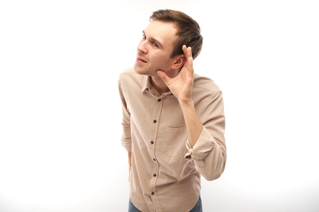 Young brunette man smiling and holding hand near ear listening carefully hearing intently isolated on white background