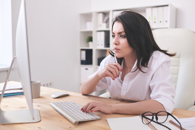 Young brunette manager focused on online project sitting at desk and reading information on computer monitor