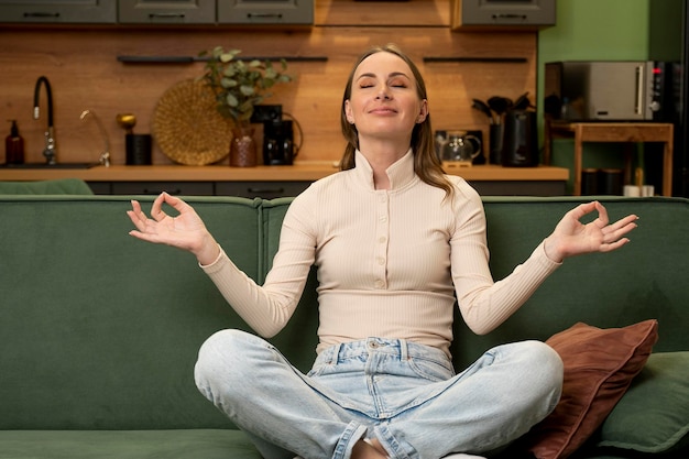 Young brunette meditates sitting in a lotus position on a green sofa in the living room at home a