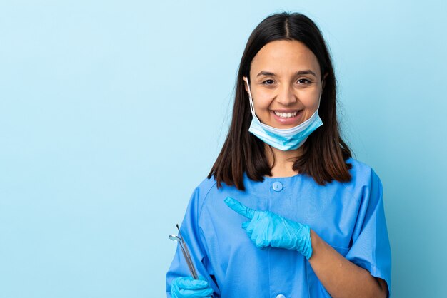 Strumenti di tenuta della giovane donna del dentista della corsa mista del brunette sopra la parete che indica il lato per presentare un prodotto