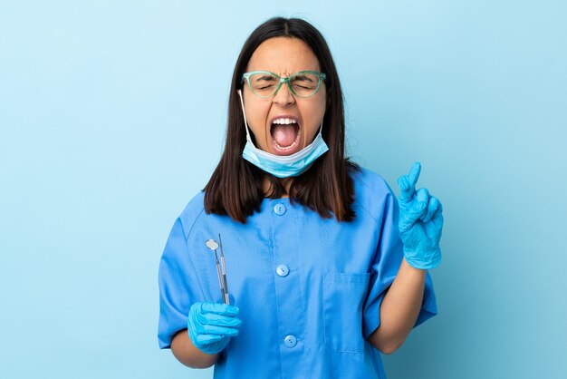 Young brunette mixed race dentist woman holding tools over wall with fingers crossing