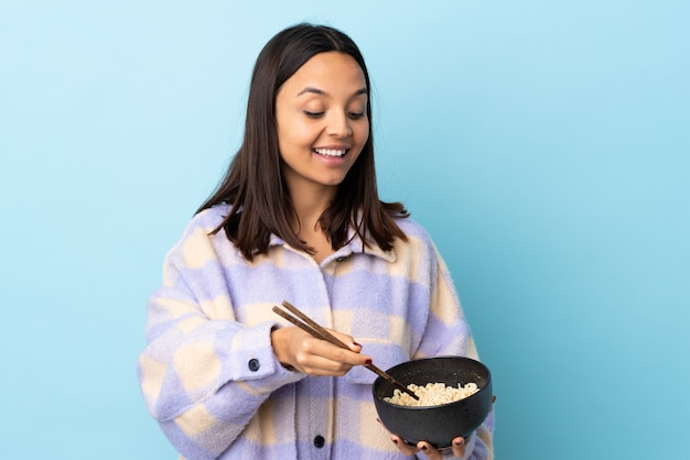 Young brunette mixed race woman over blue wall holding a bowl of noodles with chopsticks