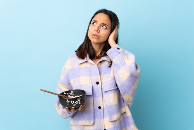 Young brunette mixed race woman holding a bowl full of noodles over blue wall frustrated and covering ears.