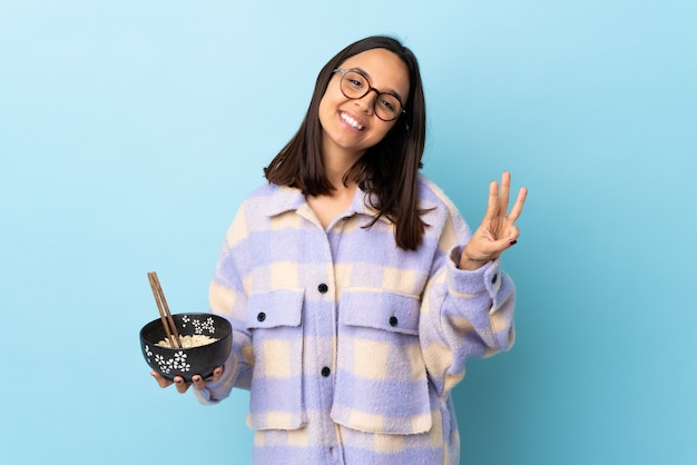 Young brunette mixed race woman holding a bowl full of noodles over blue wall happy and counting three with fingers