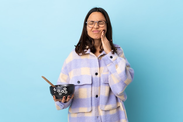 Young brunette mixed race woman holding a bowl full of noodles over blue wall with toothache.