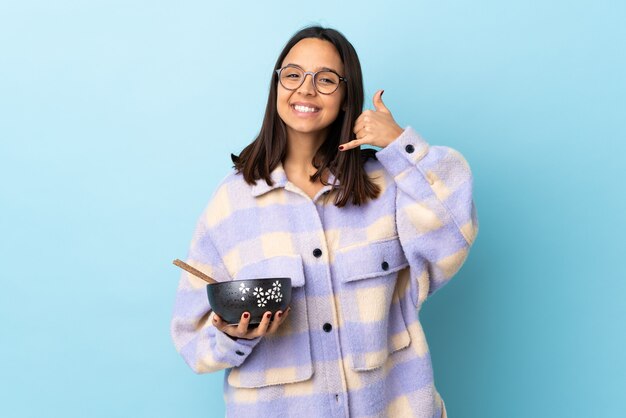 Young brunette mixed race woman holding a bowl full of noodles over isolated blue making phone gesture. Call me back sign.