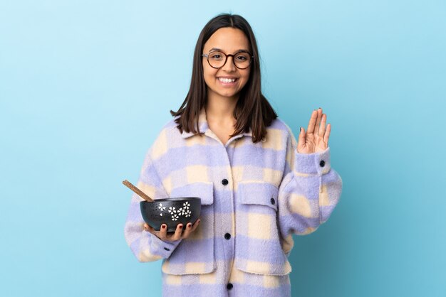 Young brunette mixed race woman holding a bowl full of noodles over isolated blue saluting