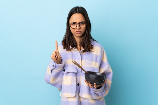 Young brunette mixed race woman holding a bowl full of noodles over isolated blue wall counting one with serious expression