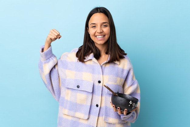 Young brunette mixed race woman holding a bowl full of noodles over isolated blue wall doing strong gesture.