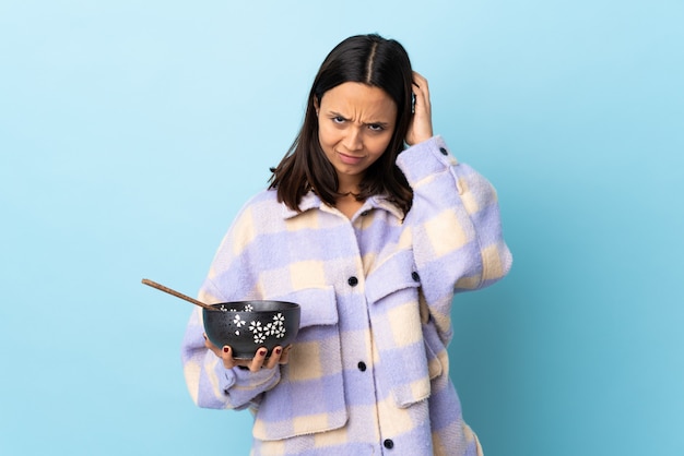 Young brunette mixed race woman holding a bowl full of noodles over isolated blue wall having doubts