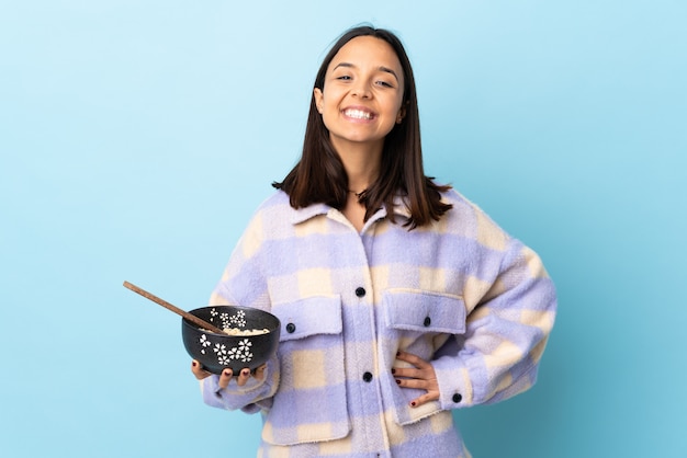 Young brunette mixed race woman holding a bowl full of noodles over isolated blue wall keeping the arms crossed in frontal position