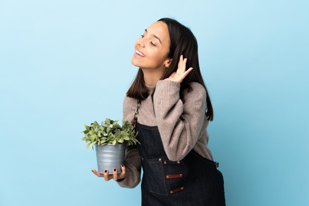 Young brunette mixed race woman holding a plant over blue wall listening to something by putting hand on the ear