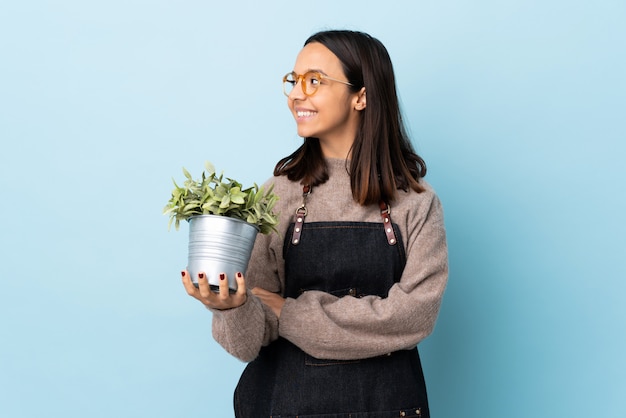 Young brunette mixed race woman holding a plant over blue wall looking side