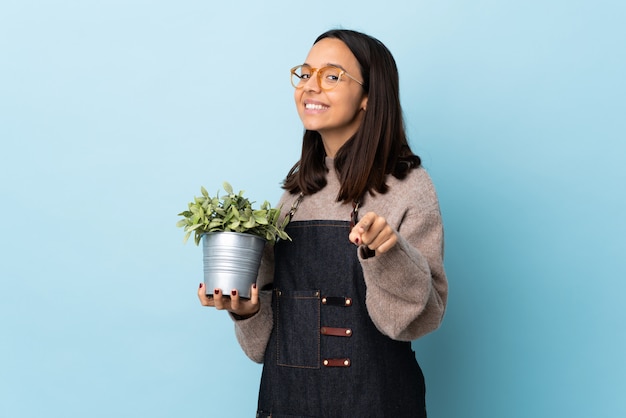 Young brunette mixed race woman holding a plant over blue wall pointing front with happy expression.