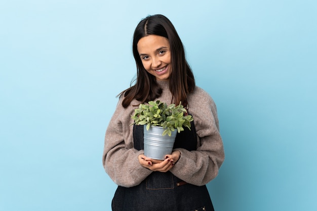 Young brunette mixed race woman holding a plant over isolated blue wall keeps palm together. Person asks for something