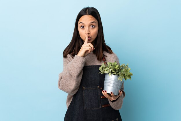 Young brunette mixed race woman holding a plant over isolated blue wall showing a sign of silence gesture putting finger in mouth