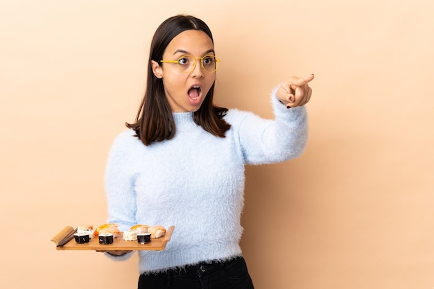 Young brunette mixed race woman holding sushi over isolated  pointing away