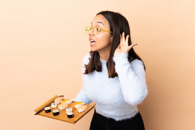 Young brunette mixed race woman holding sushi over isolated wall listening to something by putting hand on the ear