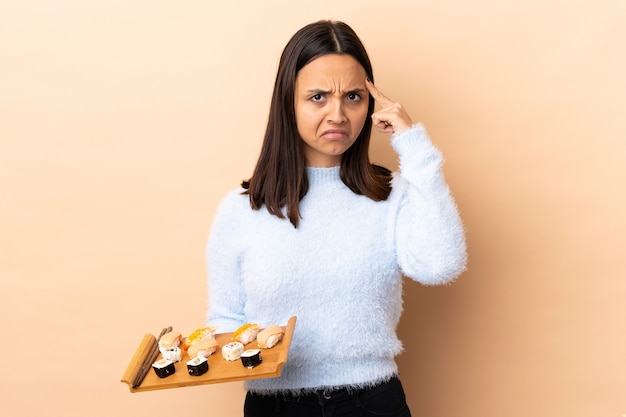 Young brunette mixed race woman holding sushi over isolated wall thinking an idea