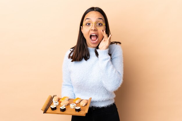 Young brunette mixed race woman holding sushi over wall shouting with mouth wide open