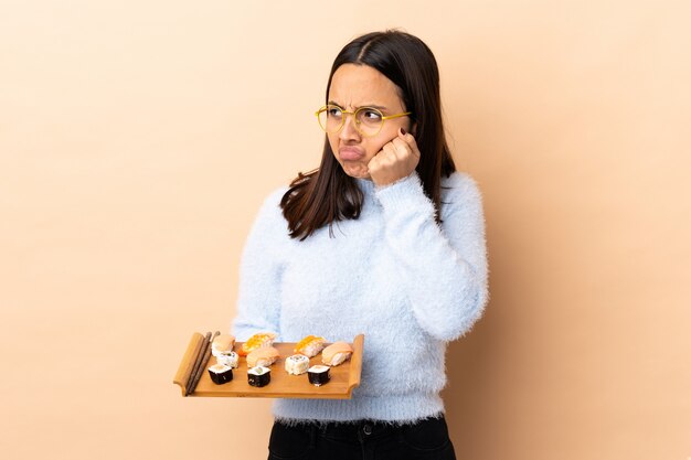 Young brunette mixed race woman holding sushi over wall with tired and bored expression