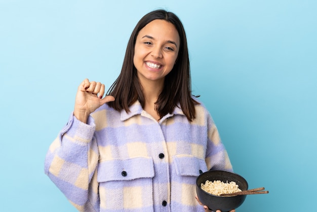 Young brunette mixed race woman over isolated blue background proud and self-satisfied while holding a bowl of noodles with chopsticks.