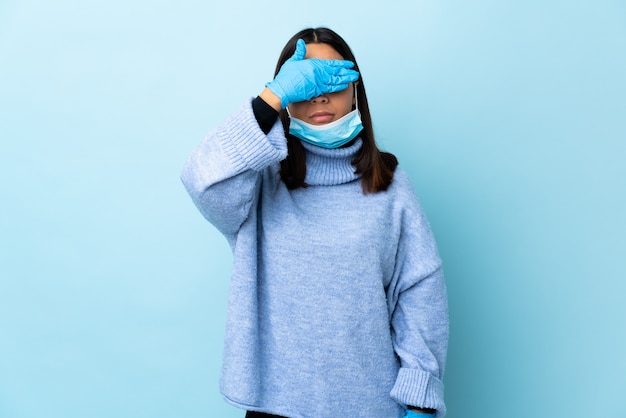 Young brunette mixed race woman protecting from the coronavirus with a mask and gloves over isolated blue wall covering eyes by hands. Do not want to see something