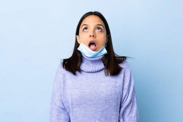 Young brunette mixed race woman protecting from the coronavirus with a mask and gloves over isolated blue wall looking up and with surprised expression