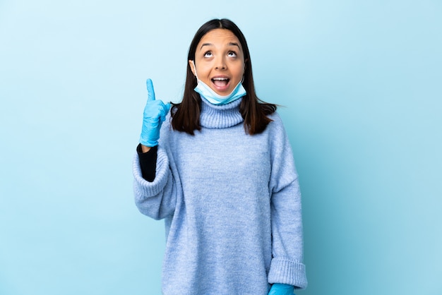 Young brunette mixed race woman protecting from the coronavirus with a mask and gloves over isolated blue wall pointing up and surprised