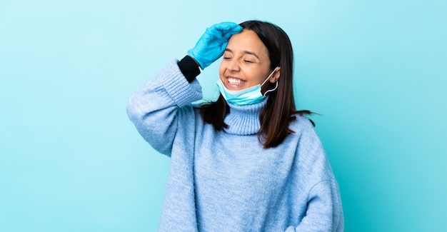 Young brunette mixed race woman protecting from the coronavirus with a mask and gloves over isolated blue wall smiling a lot