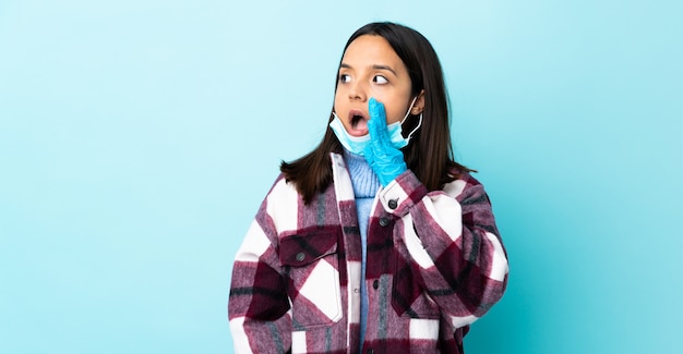 Giovane donna di razza mista bruna che protegge dal coronavirus con una maschera e guanti sul muro blu isolato sussurrando qualcosa con gesto di sorpresa mentre guarda al lato