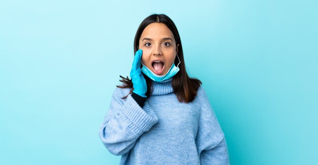 Young brunette mixed race woman protecting from the coronavirus with a mask and gloves over isolated blue wall with surprise and shocked facial expression