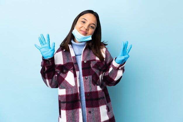 Young brunette mixed race woman protecting with a mask and gloves over blue wall counting eight with fingers