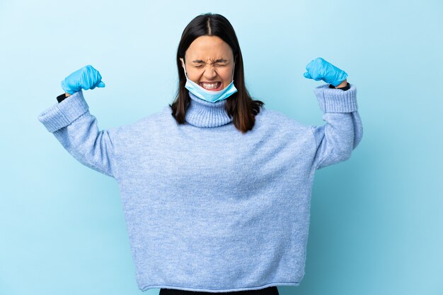 Foto giovane donna castana della corsa mista che protegge con una maschera e guanti sopra la parete blu che fa forte gesto