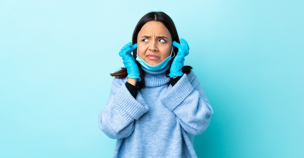 Young brunette mixed race woman protecting with a mask and gloves over blue wall frustrated and covering ears