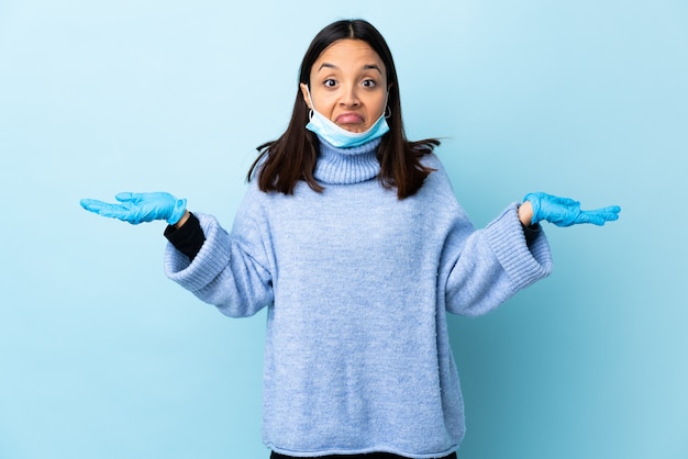 Young brunette mixed race woman protecting with a mask and gloves over blue wall having doubts while raising hands