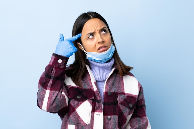Young brunette mixed race woman protecting with a mask and gloves over blue wall making the gesture of madness putting finger on the head