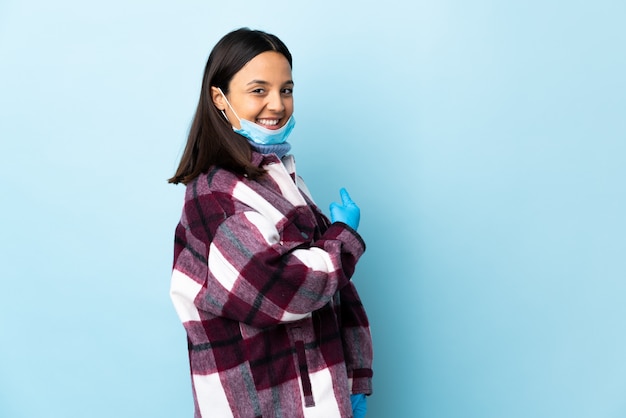 Giovane donna castana della corsa mista che protegge con una maschera e guanti sopra la parete blu che indica indietro