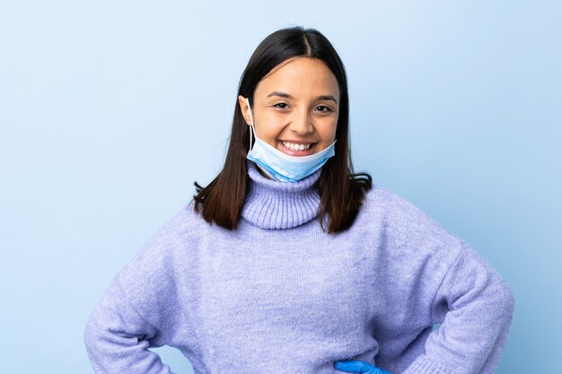 Young brunette mixed race woman protecting with a mask and gloves over blue wall posing with arms at hip and smiling