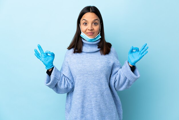 Young brunette mixed race woman protecting with a mask and gloves over blue wall in zen pose