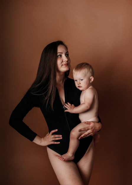 A young brunette mom in a black bodysuit holds a baby girl on a brown background with a copy of the space