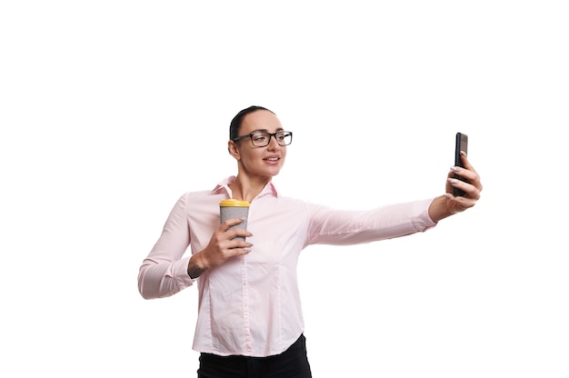 Молодая брюнетка в розовой рубашке с кружкой из переработанного картона с горячим напитком, держа мобильный телефон на расстоянии вытянутой руки.