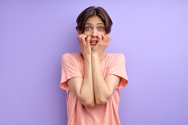Young brunette short haired woman scred by something looking at camera and touching cheeks with big eyes isolated purple background