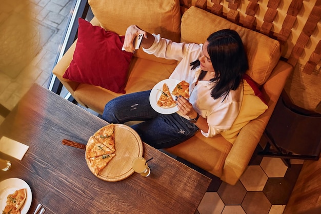 Young brunette sitting indoors with pizza and smartphone in hand.