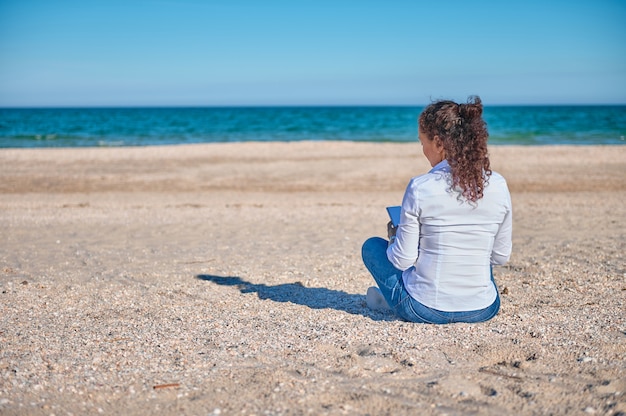 Foto giovane bruna seduta sulla sabbia di fronte al bellissimo mare blu e disegnando immagini sulla spiaggia