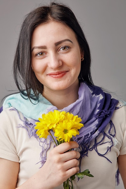 Young brunette smiling woman dressed in light shirt and scarf holding a bouquet of yellow flowers