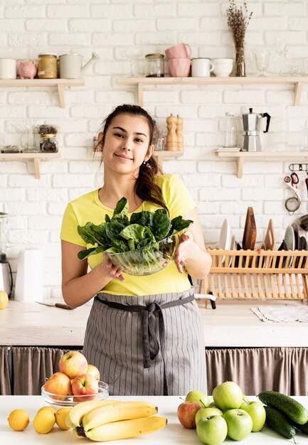 Young brunette smiling woman holding a bowl of fresh spinach in the kitchen